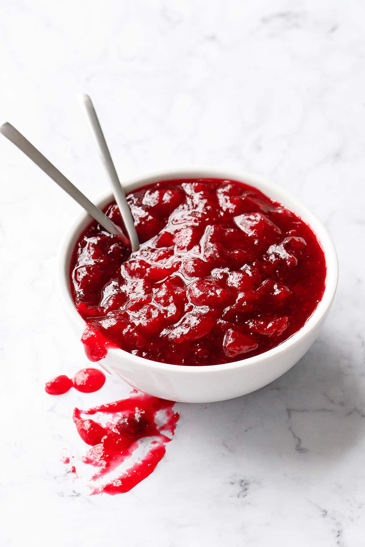 White bowl filled with a chunky cranberry sauce and two spoons on a white marble background.