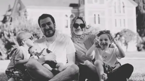 Instagram Zehra Gültekin and her husband Bilal Gültekin smile in a black and white photo seated with their children on a lawn in front of a large building. Zehra is wearing a headscarf and dark glasses, and one child - on the far left of the picture - is also wearing sunglasses