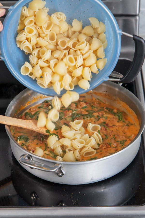 Adding cooked shellbows pasta to saucepan along with creamy sundried tomato sauce.