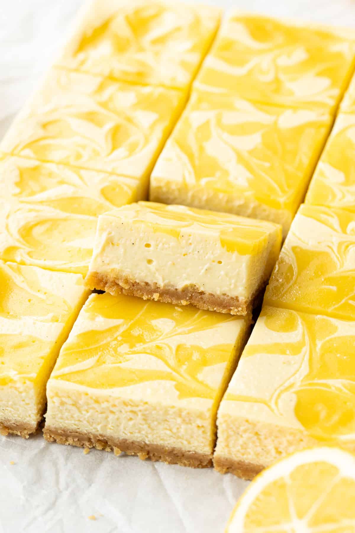 Closeup, squares of Meyer Lemon Cheesecake Bars with swirls of Lemon Curd, one piece tipped upwards to show the layers of crust and cheesecake.