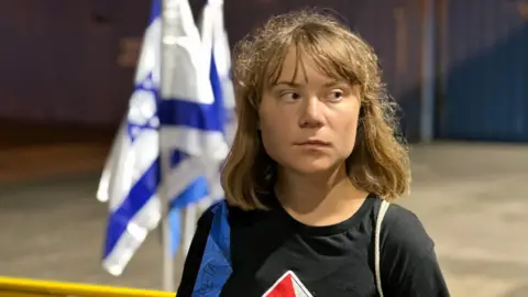 Israel Foreign Ministry Swedish activist Greta Thunberg, wearing a black T-shirt, looks to her left. Two Israeli flags can be seen in the background.