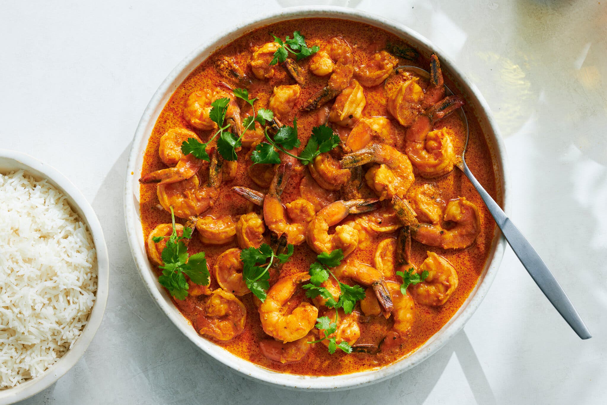 An overhead image of a red-orange shrimp curry dotted with herbs. A bowl of white rice sits to the left of the curry.