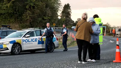 Getty Images Police and bystanders appear near a roadblock on a winding country road scattered with traffic cones.