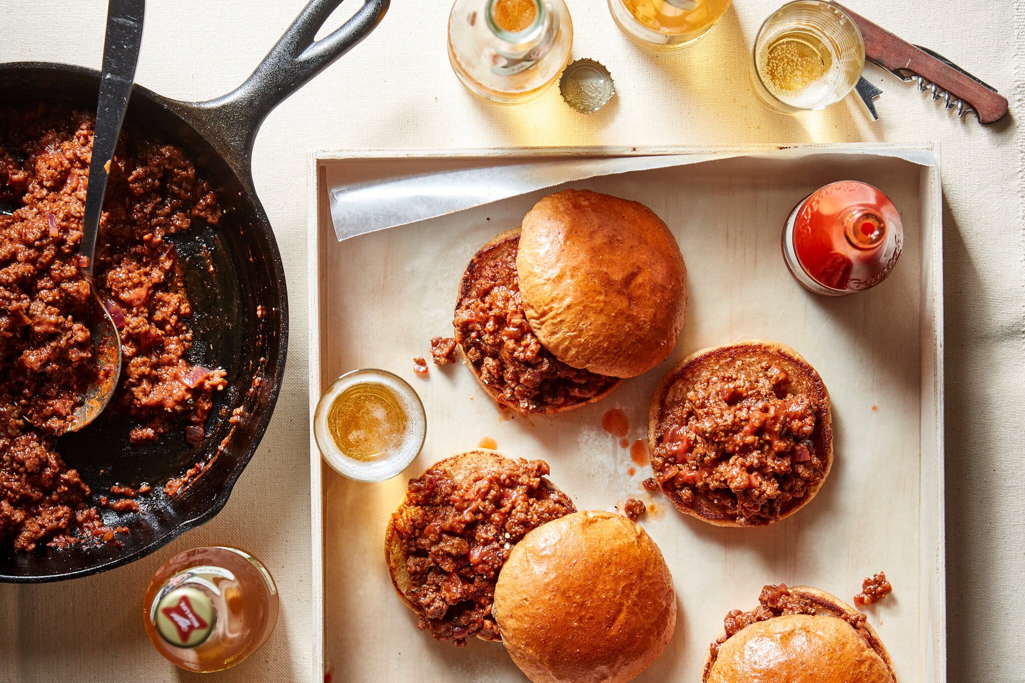 Three sloppy Joe sandwiches are arranged on a wooden tray with a glass of beer; a black cast-iron skillet containing the brown-red meat mixture sits on the left.