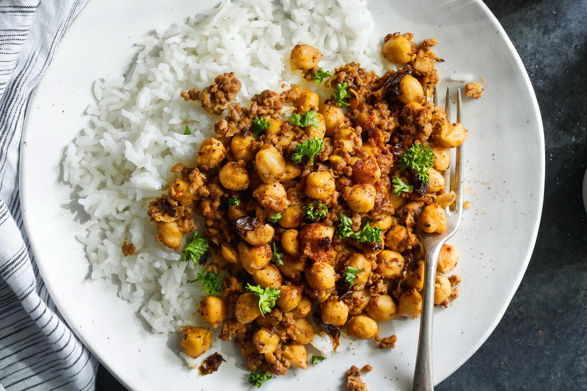 A white plate holds a generous portion of white rice next to a reddish-brown mixture of ground beef and chickpeas, with a sprinkle of green parsley; a silver fork rests on the right.