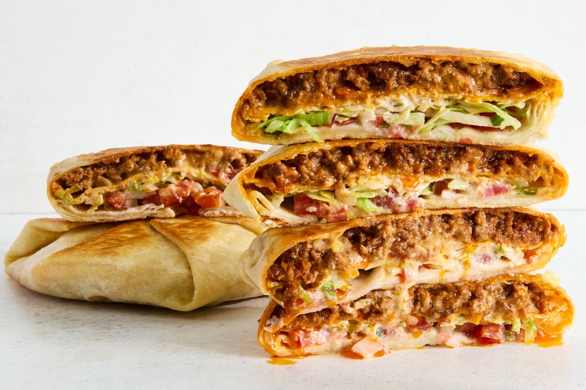 Three flattened tortilla wraps, one whole and two cut in half and stacked, reveal a cross-section of spiced ground beef, orange queso sauce, lettuce and pico de gallo inside a golden-brown tortilla.
