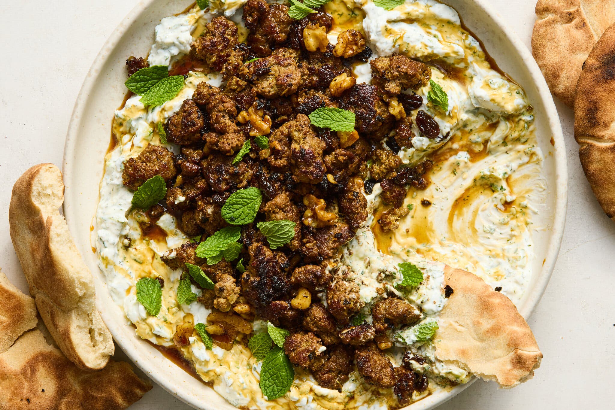 An overhead view of a spiced ground beef, raisin and walnut mixture over a thick white yogurt dip, garnished with mint leaves, served with pita bread.