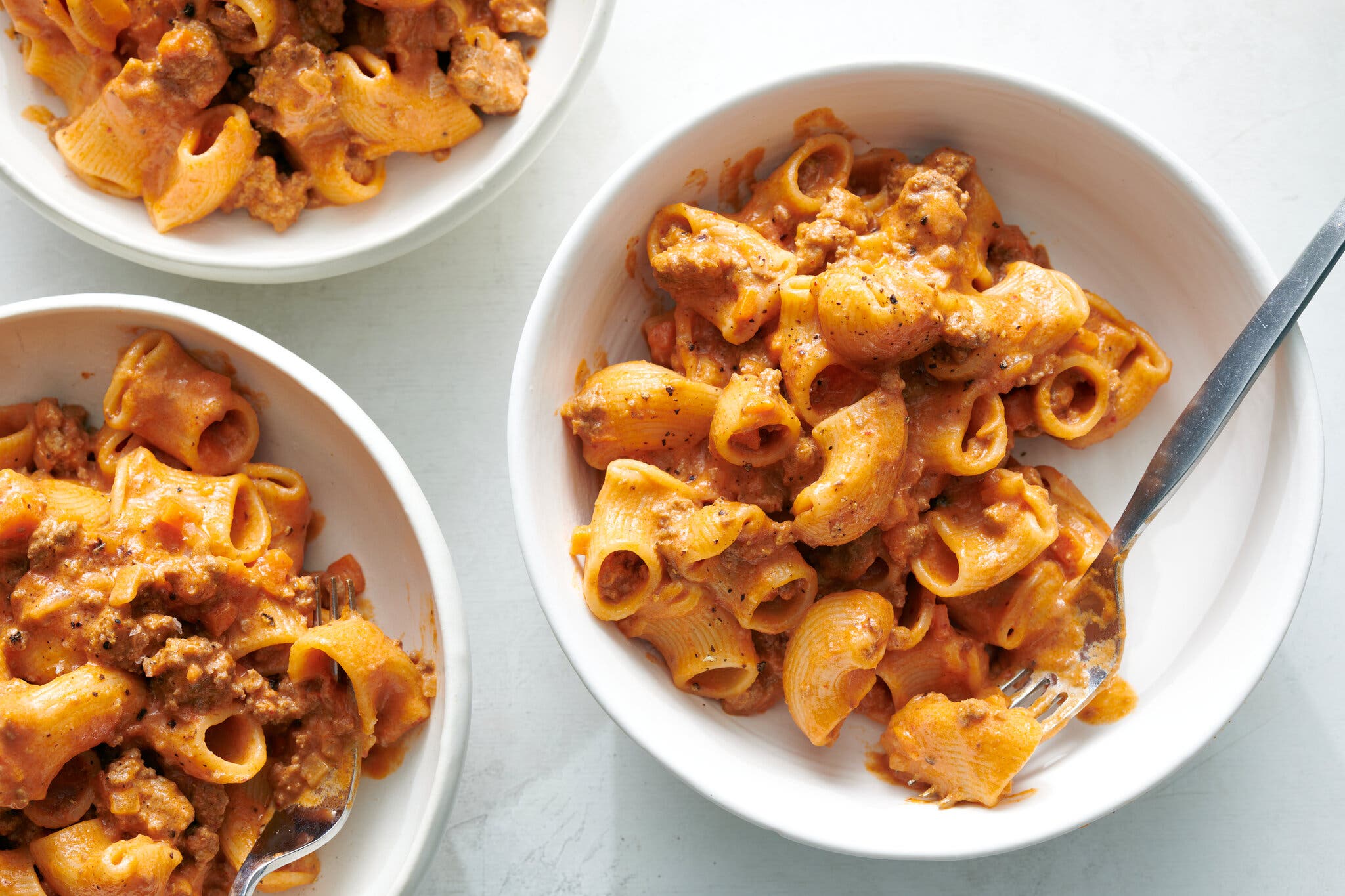 A close-up, overhead view of two bowls filled with maccheroni pasta tubes coated in a rich, reddish-orange sauce and ground beef; the bowl on the right has a silver fork resting on the side.