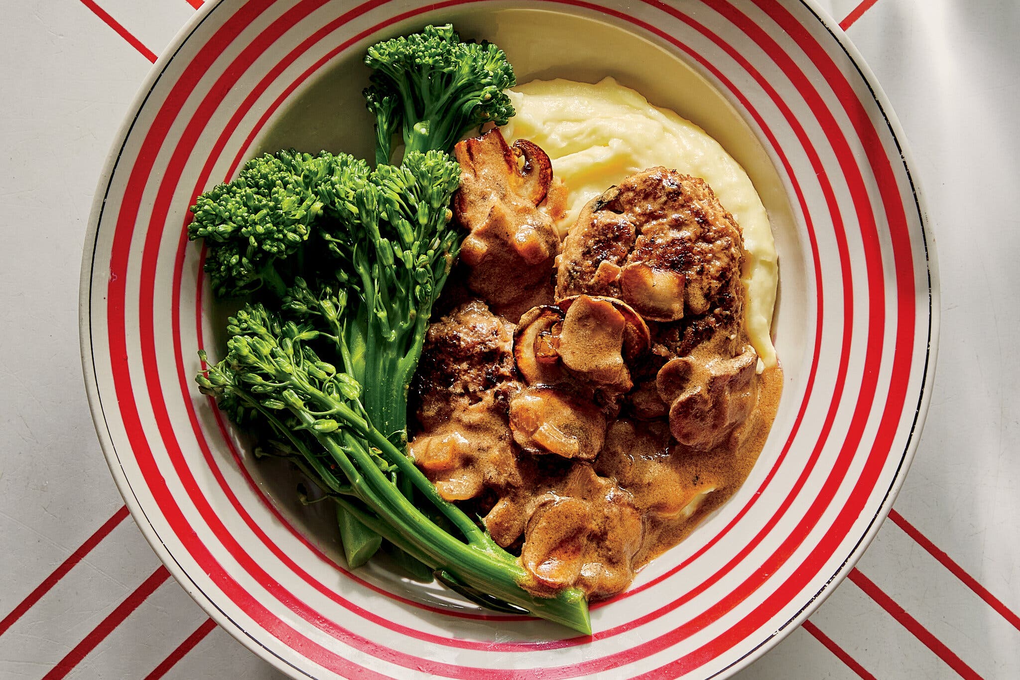 An overhead view of a dinner plate with bold red and white stripes holding a Salisbury steak covered in thick mushroom gravy, a pile of creamy mashed potatoes, and blanched green broccolini.