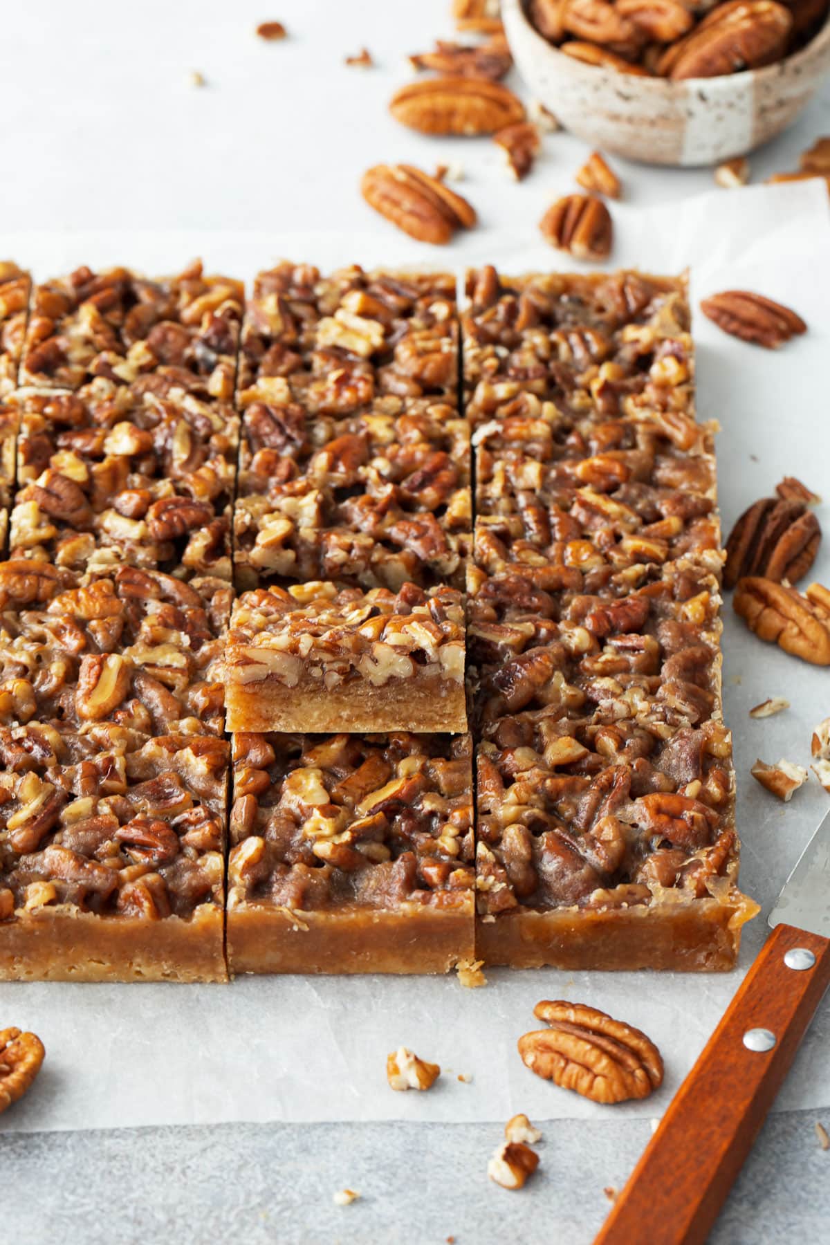 Pecan Pie Shortbread Cookie Bars cut into squares, one tilted upwards to show the layers, with a paring knife and raw pecans scattered around.